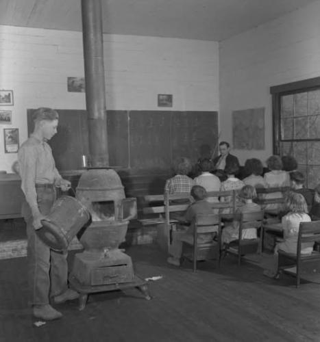 A boy pouring a bucket of coal into a stove to warm the classroom - Claiborne County, Tennessee, 1940.